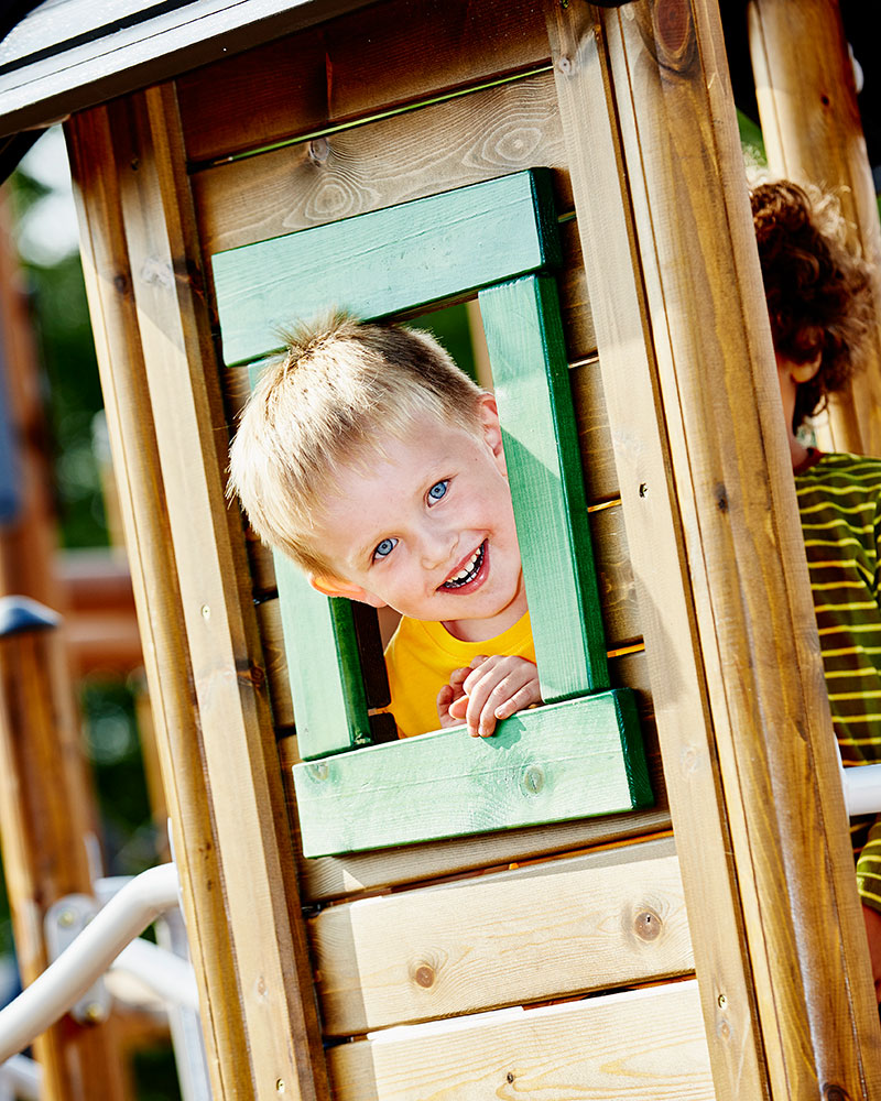 Little boy  sticks his head out from a wooden window of a wooden playground playhouse.